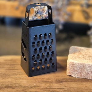 Könnte beinhalten: Eine kleine schwarze Metallreibe mit einem herzförmigen Griff. Die Reibe liegt auf einer Holzoberfläche neben einem Stück Seife.