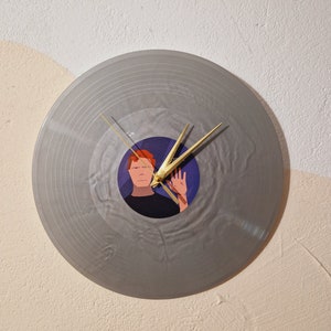 Pode incluir: Um relógio de vinil cinza com uma imagem de uma pessoa com cabelo ruivo no centro. Os ponteiros do relógio são dourados.