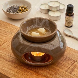 Könnte beinhalten: Ein brauner Keramik-Ölbrenner mit einem Teelicht im Inneren, das eine Schale mit Wachsschmelzen erwärmt. Daneben befinden sich eine kleine Schale mit getrocknetem Lavendel und Kamille, drei Teelichter, ein Fläschchen ätherisches Öl und ein Löffel.