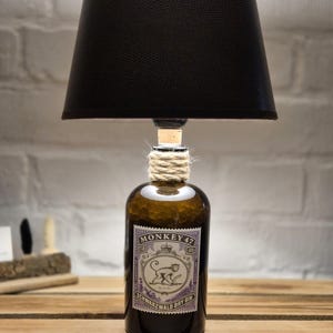 Könnte beinhalten: Eine Tischlampe aus einer wiederverwendeten Monkey 47 Gin-Flasche mit einem schwarzen Stofflampenschirm. Das Flaschenetikett zeigt einen Affen und den Text "Schwarzwald Dry Gin".