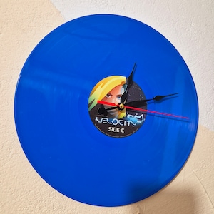 Pode incluir: Um relógio de vinil azul com um mostrador preto e vermelho. O disco tem um adesivo com o rosto de uma mulher e o texto "Velocity Side C".
