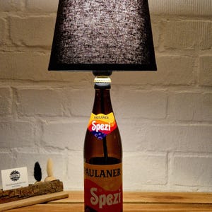 Könnte beinhalten: Eine Tischlampe mit einem braunen Paulaner Spezi Glasflaschenfuß und einem schwarzen Stofflampenschirm. Die Flasche behält ihr ursprüngliches Branding bei, einschließlich des Paulaner Spezi Logos am Flaschenhals und auf dem Hauptetikett.