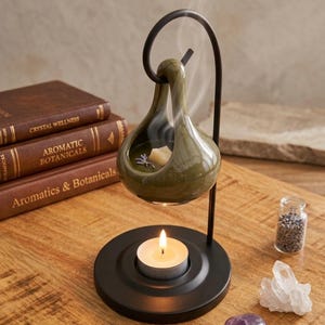 Könnte beinhalten: Ein schwarzer Metallständer hält eine glänzend olivgrüne Keramik-Duftlampe in Tropfenform. Ein brennendes Teelicht steht auf dem runden Sockel und erhitzt ein Duftwachs im Inneren, das einen dünnen Streifen weißen Rauchs aufsteigen lässt.