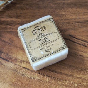 Könnte beinhalten: Ein rechteckiger Block aus festem Parfüm mit cremefarbener Basis. Die Oberseite zeigt ein Etikett im Vintage-Stil mit schwarzem Text, der PREMIUM QUALITY, MUSK LINEN und SOLID PERFUME lautet, umrahmt von dekorativen schwarzen Schnörkeln.
