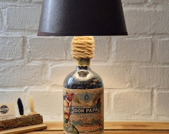 Può includere: Un'originale lampada da tavolo realizzata con una bottiglia Don Papa Baroko riciclata. La lampada presenta un paralume marrone scuro e un collo avvolto in corda. La bottiglia ha un'etichetta colorata con testo e illustrazioni.
