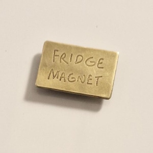 Peut inclure: Un aimant de réfrigérateur rectangulaire de couleur laiton. Les mots "FRIDGE MAGNET" sont gravés à la surface dans un style manuscrit. L'aimant est légèrement incliné, projetant une ombre douce.