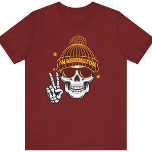 May include: Maroon t-shirt featuring a skull wearing sunglasses and a yellow beanie with the word "WASHINGTON" on it. The skull is making a peace sign with its bony hand. Yellow stars are on the shirt.