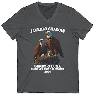 Könnte beinhalten: Dunkelgraues V-Ausschnitt-T-Shirt mit einer Grafik von zwei Weißkopfseeadlern in einem Nest mit ihren Jungen. Der Text "JACKIE & SHADOW" steht über den Adlern, und "SANDY & LUNA BIG BEAR LAKE, CALIFORNIA 2026" steht darunter.