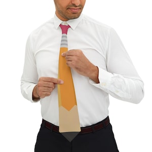 May include: A novelty tie featuring a pencil design. The tie has a pink and gray striped knot, a yellow body, and a beige and gray tip. The tie is being held by a person wearing a white dress shirt.