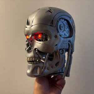 Könnte beinhalten: Ein silberfarbener, roboterartiger Totenkopf mit komplexen mechanischen Details, freiliegenden zahnradartigen Strukturen an der Schläfe und leuchtend roten LED-Augen. Die metallische Oberfläche ist glatt und weist sichtbare Gelenke auf.