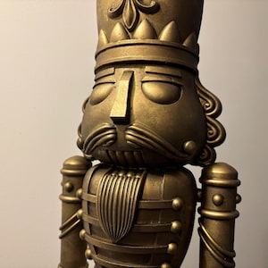 Puede incluir: Una figura de cascanueces de color dorado con una corona y una cara detallada. El cascanueces lleva un uniforme dorado con detalles intrincados.