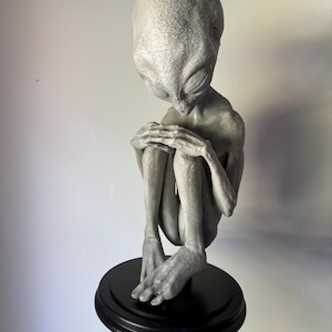Puede incluir: Escultura de un alienígena gris con una cabeza grande, sentado con los brazos alrededor de las rodillas. El alienígena está montado sobre una base redonda y negra. La escultura es detallada y parece estar hecha de un material similar a la piedra.