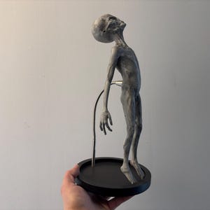 Op de afbeelding: Een grijze alien figuur met een slank lichaam en een groot hoofd, gemonteerd op een zwarte cirkelvormige basis. De alien poseert met zijn hoofd achterover gekanteld en wordt ondersteund door een dunne metalen staaf.