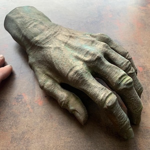 May include: A detailed, realistic sculpture of a human hand, cast in a green-brown color. The hand is posed with the fingers slightly curled, as if reaching out or grasping.