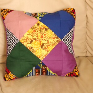 May include: A colorful patchwork pillow with a diamond pattern. The pillow is made of six different fabrics in shades of pink, yellow, green, blue, purple, and black. The pillow is sitting on a beige couch.