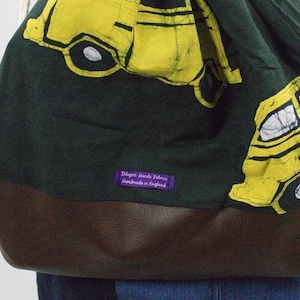 May include: A green and brown drawstring bag with a yellow and white bus pattern. The bag has a purple label that reads "Delgont Hand Fabrics handmade in England".