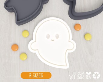 May include: A white ghost-shaped cookie cutter with a simple face design featuring two oval eyes and a small curved mouth. The cutter is surrounded by small yellow and orange round candies, with dark gray cutter parts visible at the top.