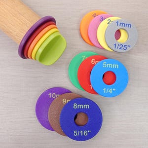 May include: A wooden rolling pin with a series of coloured silicone rings and a set of multi-coloured rolling pin guides. The guides are labelled with measurements in inches and millimetres, including 1/25", 1/4", and 5/16".