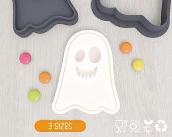 May include: A white ghost-shaped cookie cutter with a cutout face featuring two oval eyes and a jagged mouth. The set includes two dark gray outer frames and several small, round, multicolored candies scattered around the white cutter.