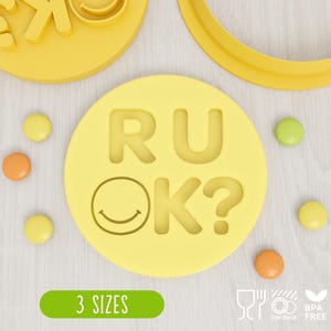 May include: A yellow circular cookie cutter mould featuring the embossed text RU OK? with a smiley face replacing the letter O. The set includes three distinct sizes of the mould, accompanied by small round sweets scattered around the central piece.