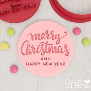 Może przedstawiać: Jasnoróżowa okrągła pieczątka do ciastek z wytłoczonym napisem merry Christmas AND HAPPY NEW YEAR w połączeniu pisma odręcznego i drukowanego, w towarzystwie małych żółtych i różowych okrągłych cukierków na jasnym tle.