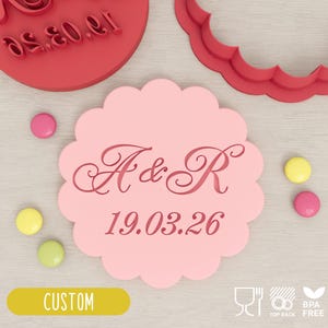 May include: A pink scalloped-edge cookie cutter with the initials "A & R" and the date "19.03.26" in a decorative font. A red circular stamp and a red scalloped-edge cutter are also visible. The word "CUSTOM" is on a yellow label.