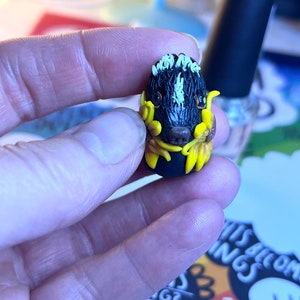 May include: A small black and white skunk figurine with yellow flowers around it. The figurine is made of polymer clay and is about 5 cm tall.