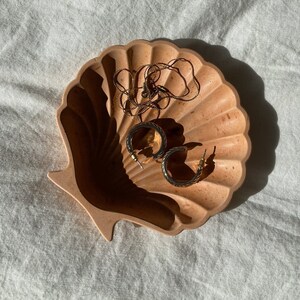Peut inclure: Un plat en forme de coquille de couleur beige clair avec un collier en chaîne dorée et deux boucles d'oreilles créoles dorées à l'intérieur.