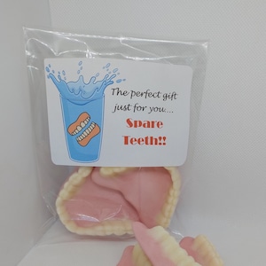 May include: A clear plastic bag containing three sets of pink and white plastic dentures. The bag has a card attached to it that says "The perfect gift just for you... Spare Teeth!" with a cartoon image of a glass of water with dentures in it.