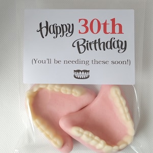 May include: A clear plastic bag with a white card that says "Happy 30th Birthday (You'll be needing these soon!)" and a drawing of dentures. The bag contains two pink and white gummy dentures.