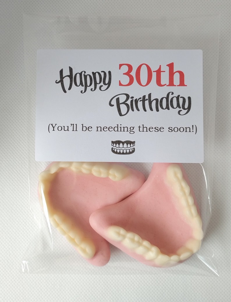May include: A clear plastic bag with a white card that says "Happy 30th Birthday (You'll be needing these soon!)" and a drawing of dentures. The bag contains two pink and white gummy dentures.