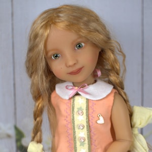 Könnte beinhalten: Eine handgefertigte Puppe mit langem, gewelltem blondem Haar und grünen Augen. Die Puppe trägt ein pfirsichfarbenes Kleid mit weißem Kragen und einer rosa Schleife. Das Kleid ist mit dekorativen Nähten und einer herzförmigen Anstecknadel verziert. Das Haar der Puppe ist zu Zöpfen mit gelben Bändern geflochten.