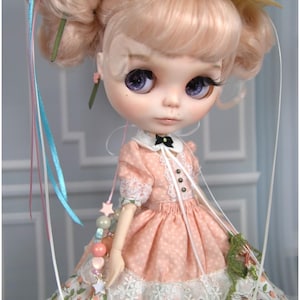 Può includere: Una bambola con capelli rosa e grandi occhi viola, che indossa un vestito color pesca con finiture in pizzo bianco e una gonna floreale. La bambola ha un cappello di paglia con nastri e tiene in mano un piccolo bouquet. Malena Art Dolls.