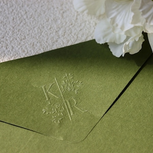 Könnte beinhalten: Nahaufnahme eines grünen Umschlags mit geprägten Initialen "KR" und floralem Design. Eine weiße Blume und strukturiertes weißes Papier befinden sich im Hintergrund. Der Umschlag ist geöffnet und zeigt ein schwarzes Innere.