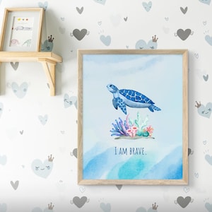 May include: A framed wall art print featuring a watercolor illustration of a blue sea turtle swimming above colorful coral reefs. The background is a soft light blue with a watercolor wash effect. The text I AM BRAVE is printed in blue.