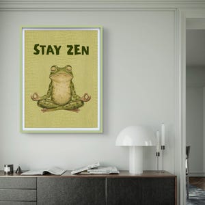 Könnte beinhalten: Gerahmter Druck mit einer grünen Frosch in Yoga-Pose und den Worten "STAY ZEN". Der Frosch befindet sich auf einem strukturierten, hellgrünen Hintergrund, der von einem hellgrünen Rahmen umgeben ist. Der Druck wird an einer Wand angezeigt.