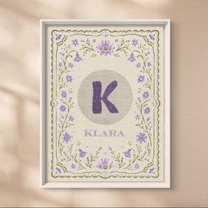 Könnte beinhalten: Ein gerahmter rechteckiger Druck auf hellbeigem, strukturiertem Hintergrund mit einem großen violetten Buchstaben K, der in einem gepunkteten Kreis zentriert ist. Unter dem Buchstaben steht der Name KLARA in Violett. Umrandet von violetten Blumenranken.