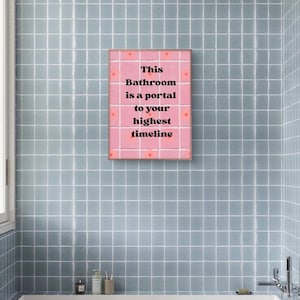Könnte beinhalten: Ein rosa gerahmter Druck mit dem Text "This Bathroom is a portal to your highest timeline" hängt an einer blau gefliesten Wand. Das Badezimmer hat eine weiße Badewanne, ein Fenster und Toilettenartikel.