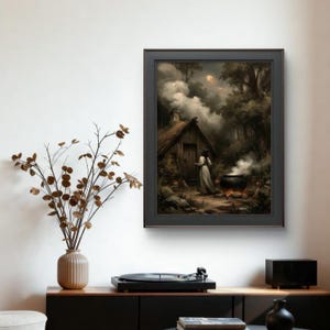 Könnte beinhalten: Ein gerahmter Wandkunstdruck, der eine dunkle, atmosphärische Waldszene mit einer kleinen Holzhütte, einer in ein langes Gewand gekleideten Figur am Eingang und einem dampfenden schwarzen Kessel über einem Feuer unter einem bewölkten Nachthimmel zeigt.