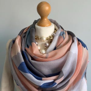 May include: A soft, pink, blue, and gray patterned scarf with a white background. The scarf is folded and draped around a wooden mannequin.