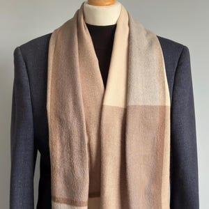 Könnte beinhalten: Ein beigefarbener und brauner Schal mit Karomuster, drapiert über einem dunkelblauen Blazer. Der Schal hat ein rechteckiges Muster mit verschiedenen Beige- und Brauntönen. Der Schal wird auf einer Schaufensterpuppe präsentiert.