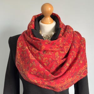 May include: A vibrant red scarf with a gold floral pattern is displayed on a mannequin. The scarf is wrapped around the neck and shoulders, over a black coat. A gold and pearl necklace is visible.