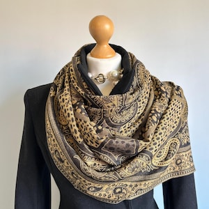 May include: A patterned scarf in black and tan, featuring paisley and leopard print motifs. The scarf is displayed on a mannequin, highlighting its design. A black jacket and a pearl necklace are also visible in the image.