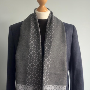 Könnte beinhalten: Ein grauer Schal mit einem weißen geometrischen Muster. Der Schal ist gefaltet und über eine Schaufensterpuppe gelegt, die einen dunkelblauen Blazer trägt.