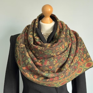 May include: A brown scarf with a floral pattern in red, green, and yellow is draped around a mannequin's neck. The scarf has a detailed border and is paired with a black jacket.
