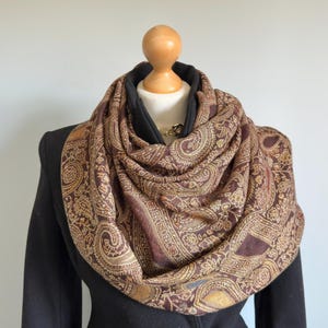 Può includere: Sciarpa a infinito con motivo paisley marrone e oro. La sciarpa è drappeggiata su un cappotto nero su un manichino. La sciarpa presenta un motivo floreale ripetuto con una combinazione di colori marrone scuro e oro. La sciarpa è realizzata in tessuto morbido e fluido.