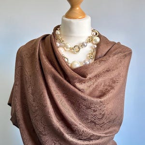 Pode incluir: Um xale castanho com um padrão paisley tonal drapeado sobre um manequim. O xale é complementado por um colar de várias camadas composto por elos de metal dourado e grandes pérolas artificiais de cor creme de vários tamanhos.