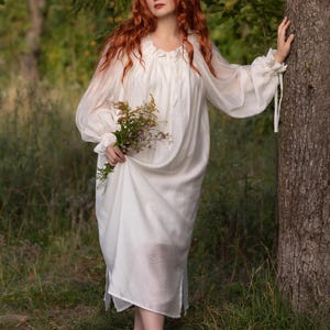 May include: A long-sleeved white dress featuring a gathered neckline with a tie closure, loose flowing sleeves with ruffled cuffs, and a relaxed silhouette. The garment is made of a lightweight, semi-sheer fabric that drapes down to the mid-calf.