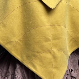 May include: A mustard yellow triangular cape featuring dark red topstitching that creates a leaf-like vein pattern across the fabric. The garment is layered over a brown skirt with a ruffled hemline, showcasing a distinct pointed silhouette.
