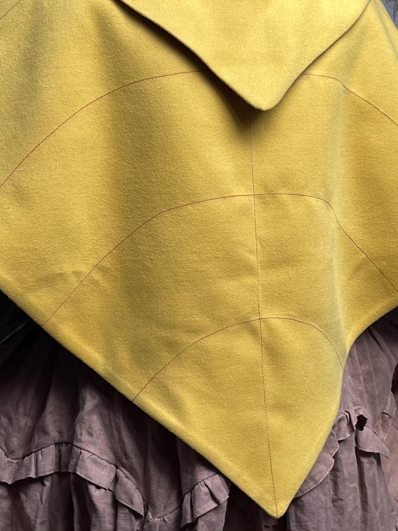 May include: A mustard yellow triangular cape featuring dark red topstitching that creates a leaf-like vein pattern across the fabric. The garment is layered over a brown skirt with a ruffled hemline, showcasing a distinct pointed silhouette.